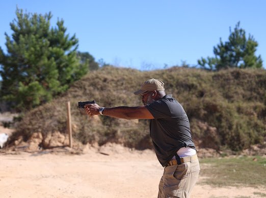A man shooting a handgun on a shooting range with trees and dirt mounds in the background.