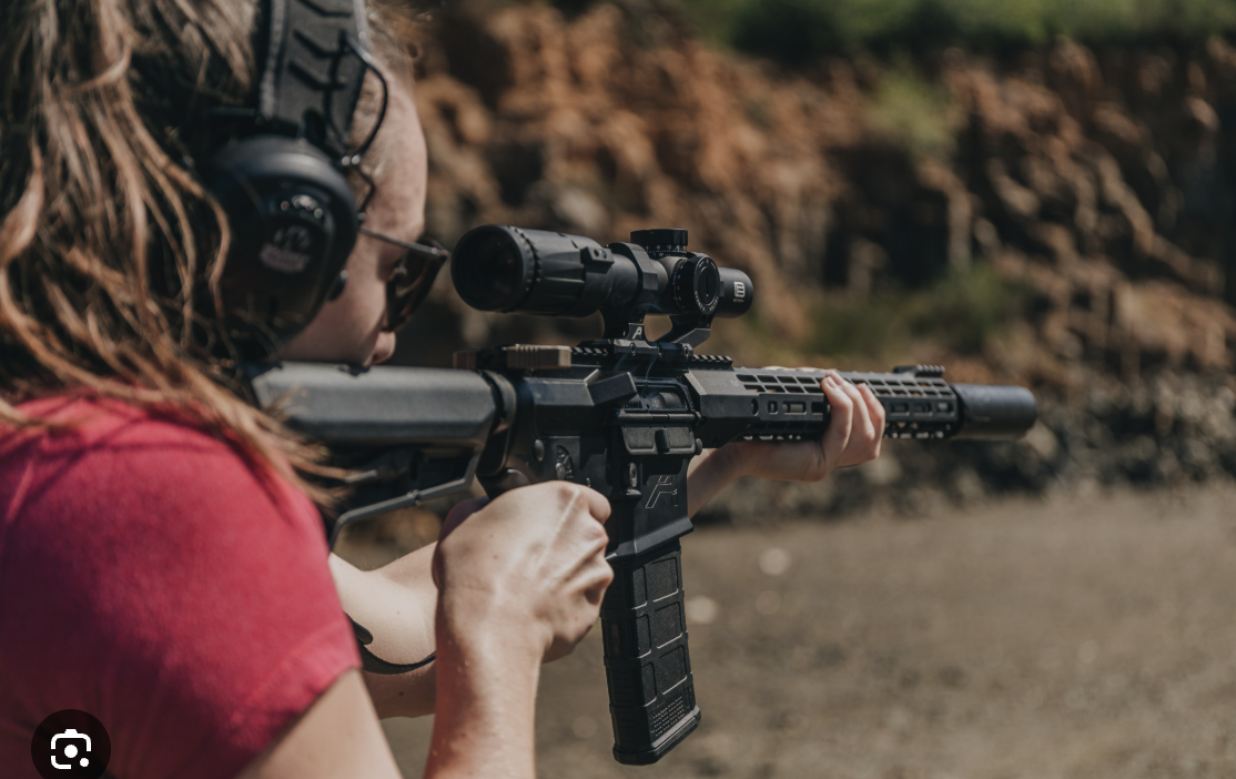 Woman in red shirt aiming a rifle with a scope outdoors.