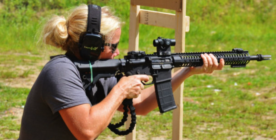 Woman with blonde hair, wearing black headphones and sunglasses, aiming a rifle at a shooting range with a wooden structure in the background.
