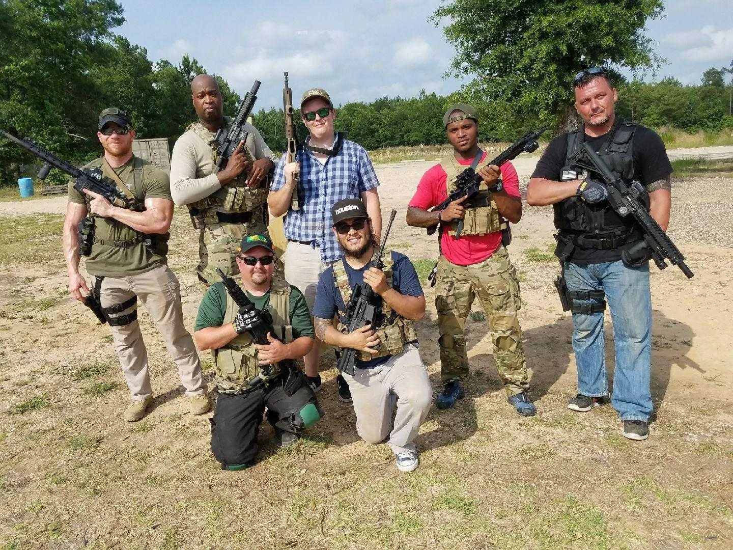 Group of seven men outdoors holding rifles on a sunny day, some wearing camouflage and tactical gear, in an open field with trees in the background.