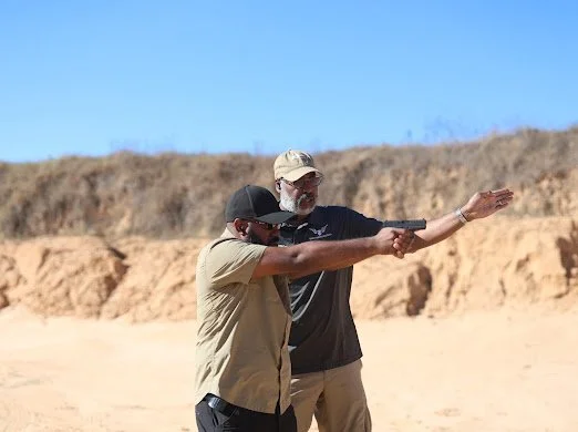 Two men outdoors on a sunny day, one guiding the other in aiming a firearm.