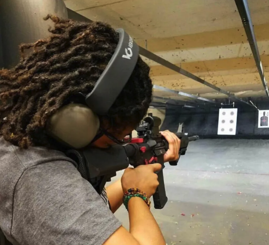 A person with dreadlocks wearing hearing protection and a gray shirt, aiming a rifle in an indoor shooting range with target targets visible in the background.