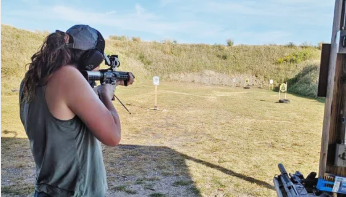 Woman in sleeveless shirt and headphones shooting a rifle at outdoor shooting range.