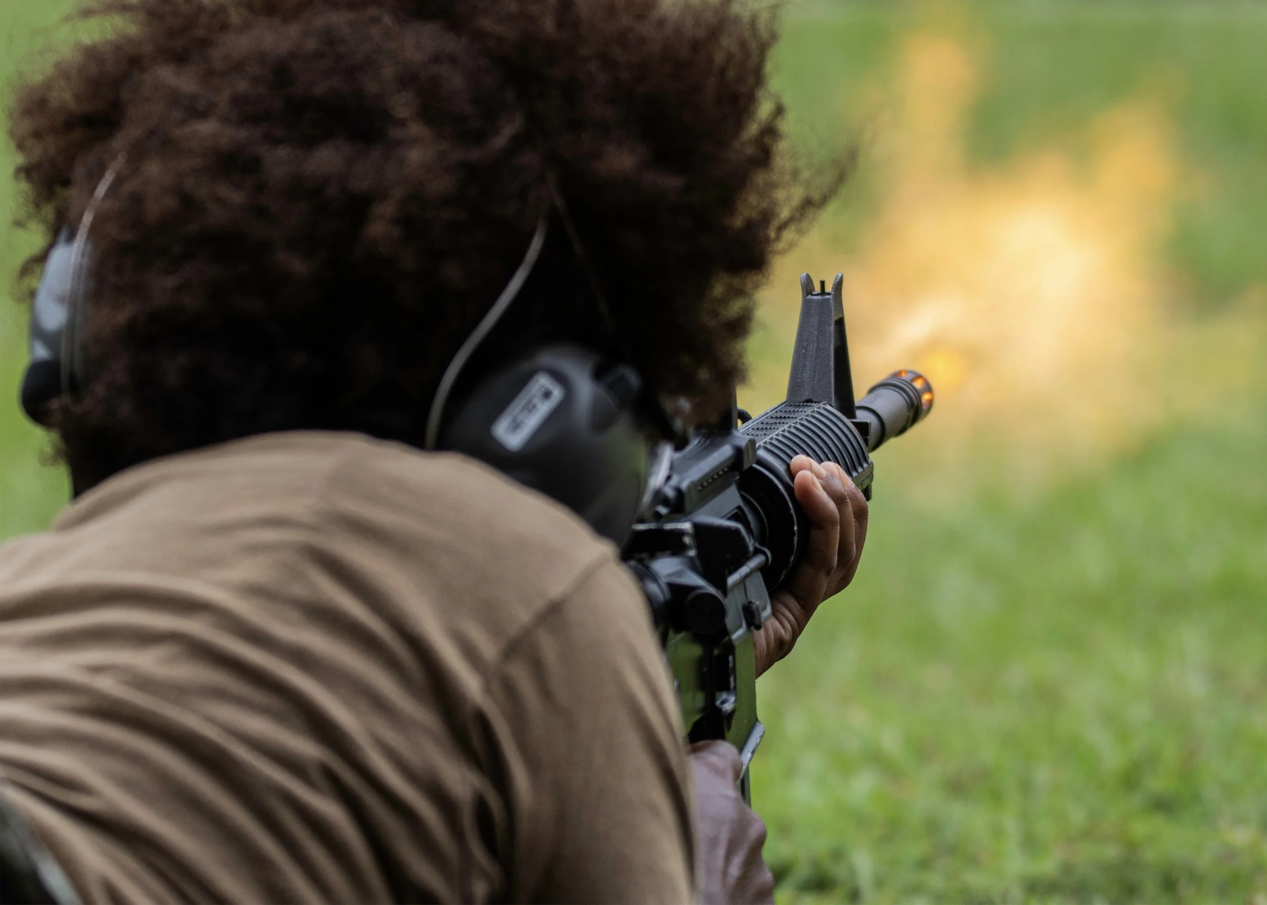 Person shooting a rifle outdoors with a green and yellow blurred background.