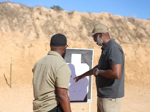 Two men looking at a silhouette target on a shooting range, with dirt mounds in the background.