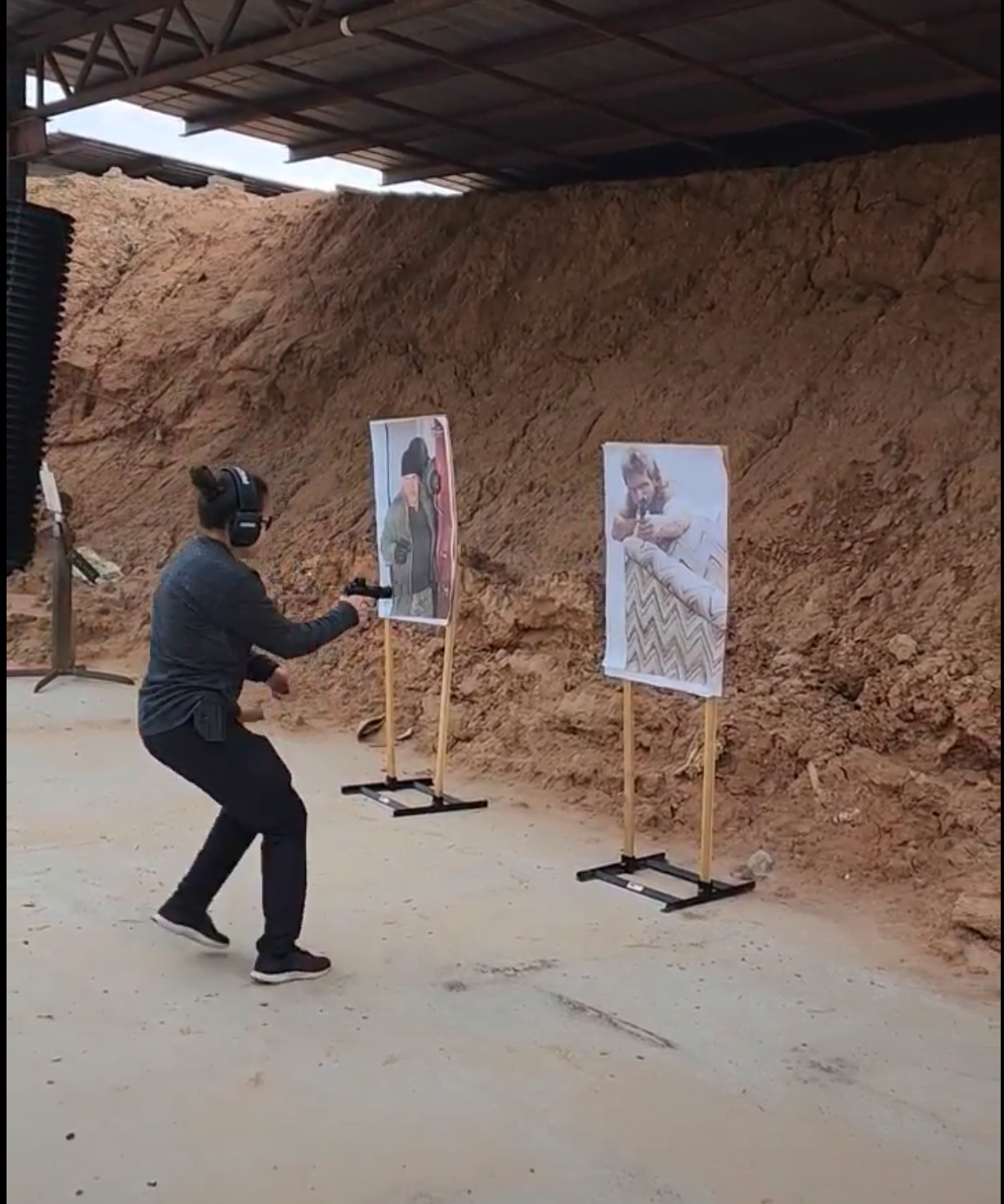 A person wearing headphones and black clothing posing with a gun at a shooting range with two posters of women in the background.