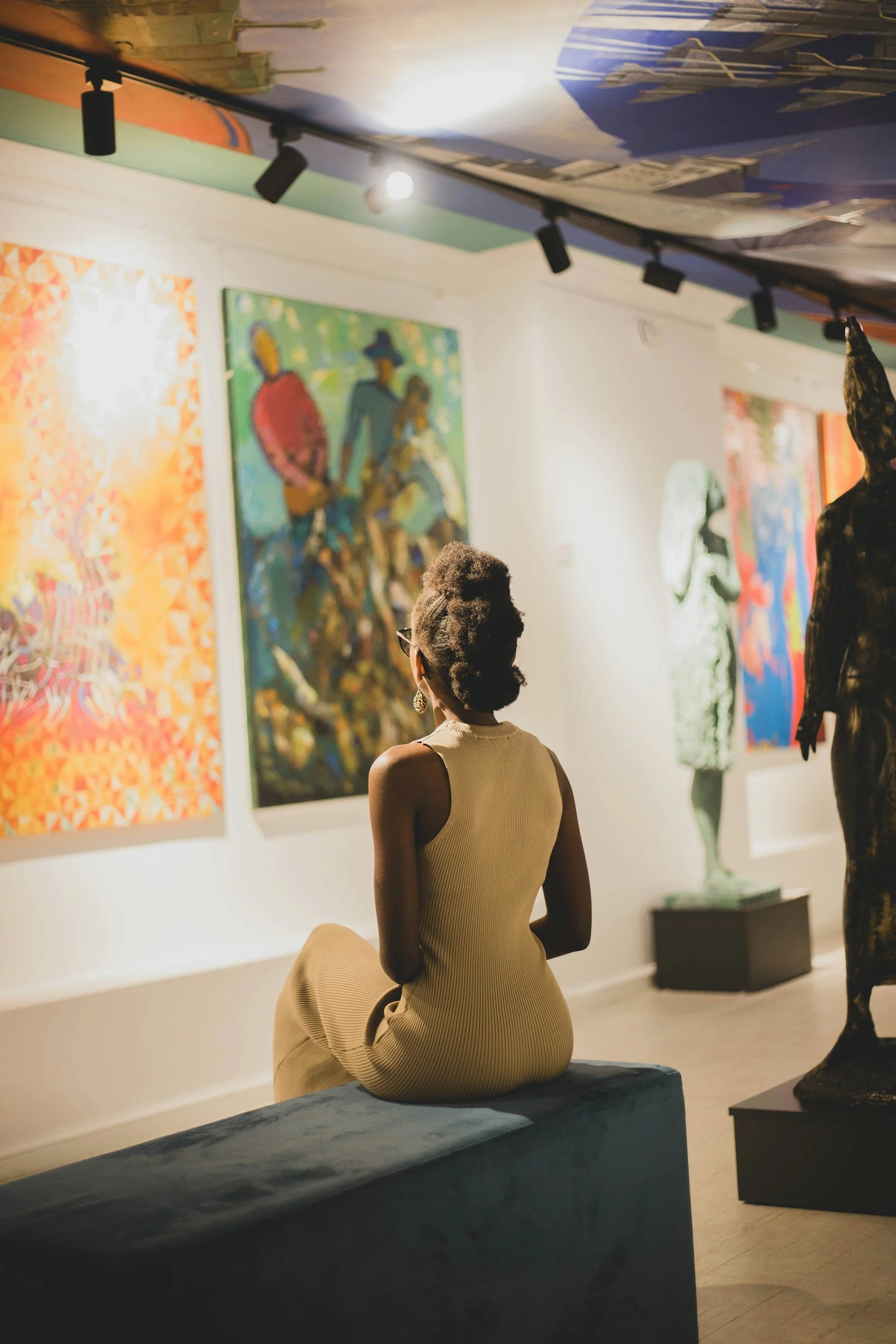 A woman wearing a beige dress is sitting on a blue ottoman in an art gallery, observing colorful paintings and sculptures on the walls and pedestal.