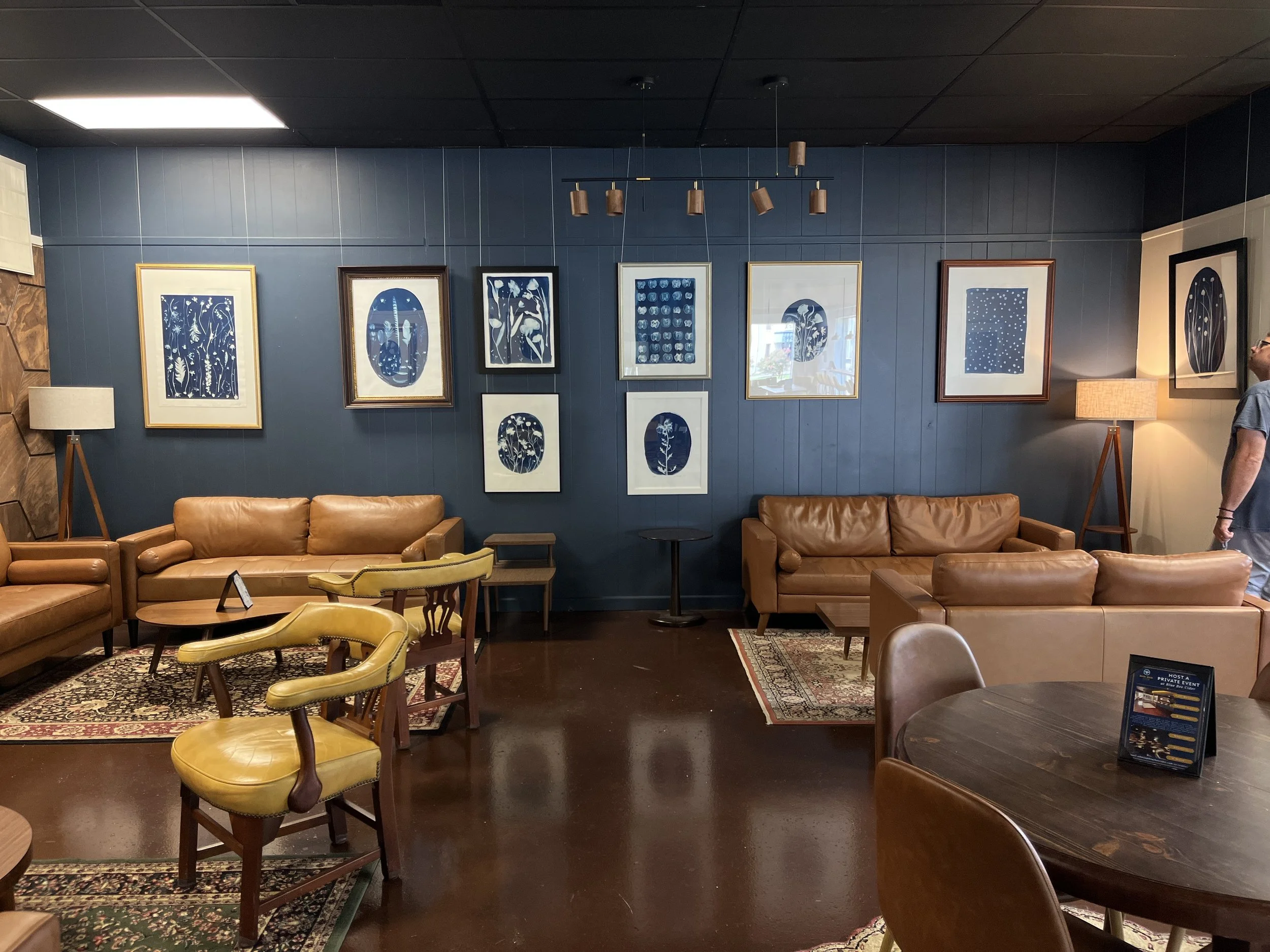 Interior of a lounge or cafe with leather sofas, wooden chairs, lamps, and framed artwork on a blue wall.