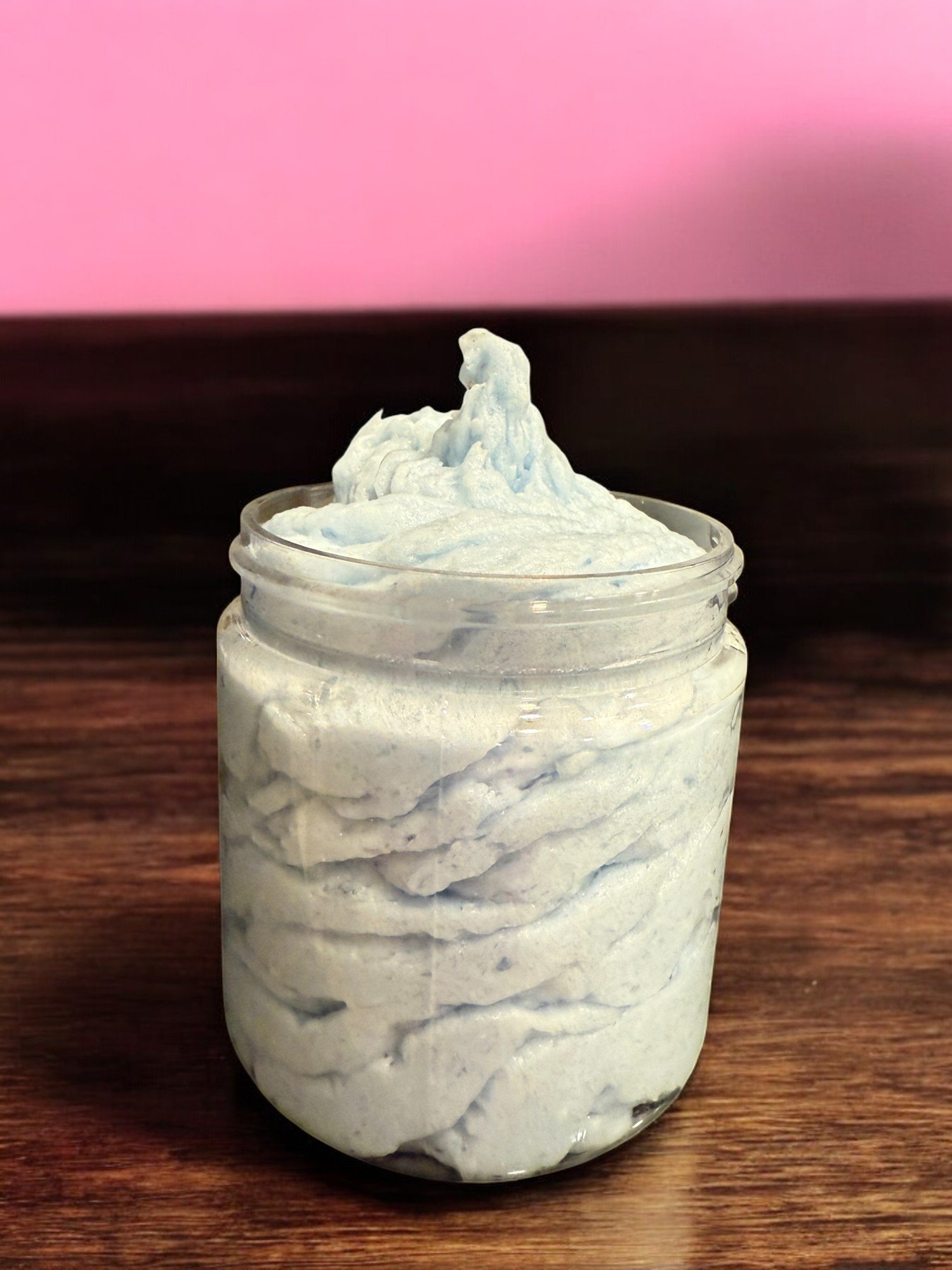 A glass jar filled with a white, fluffy, whipped substance, sitting on a wooden surface with a pink and black background.