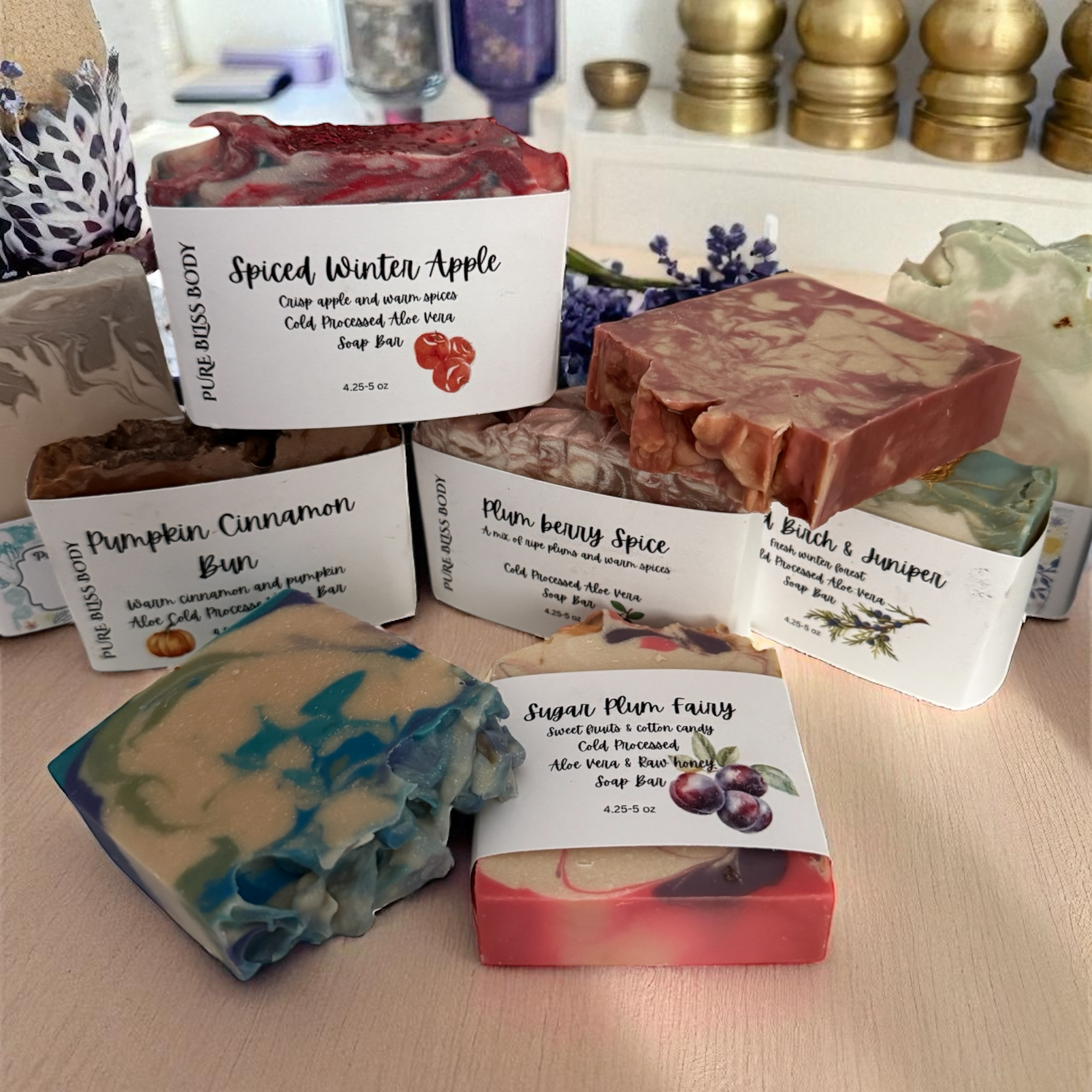 A collection of colorful handcrafted soap bars with various labels describing their scents and ingredients, including 'Spiced Winter Apple,' 'Pumpkin Cinnamon Bun,' 'Plum Berry Spice,' 'Birch & Juniper,' and 'Sugar Plum Fairy.'