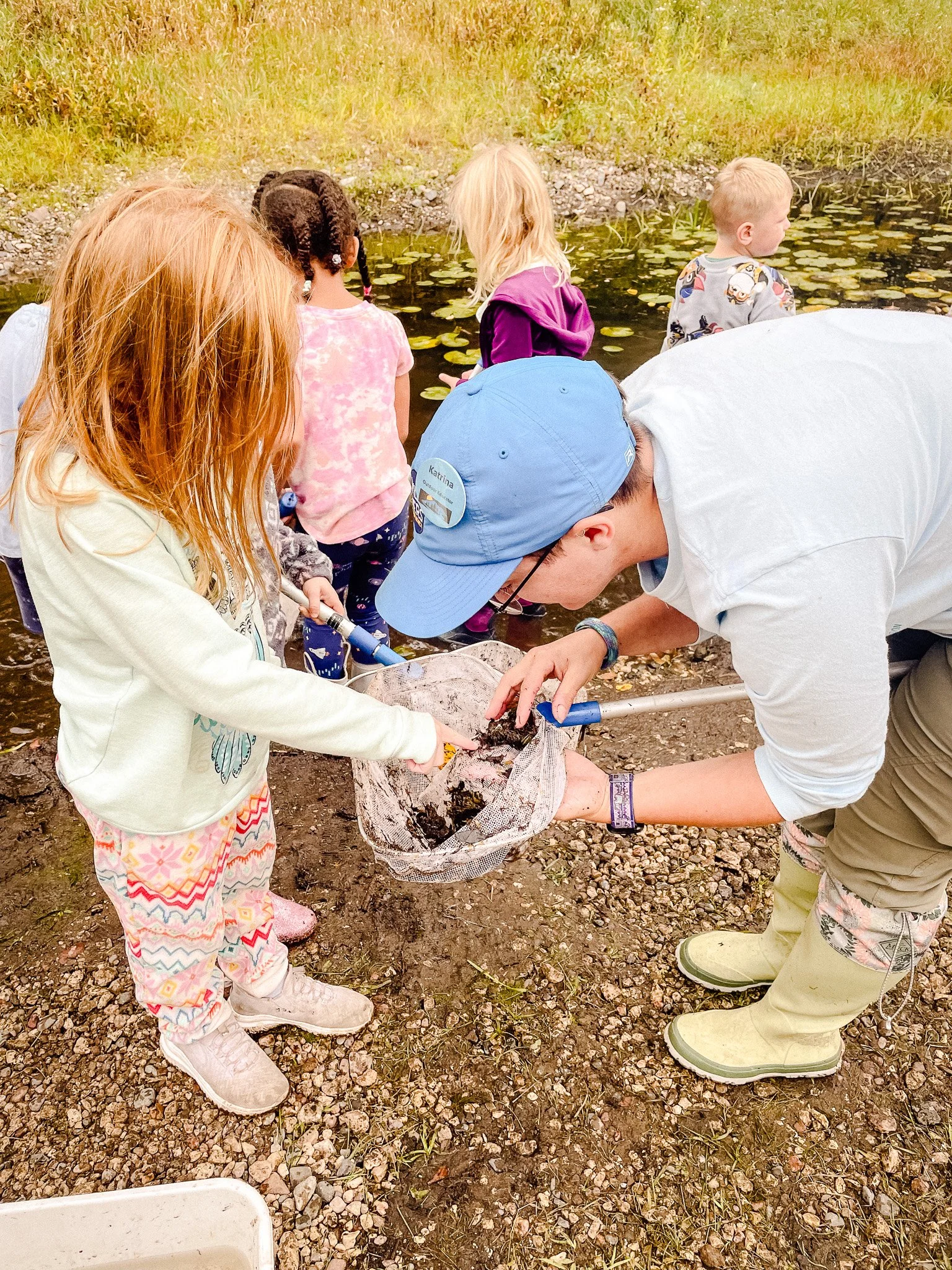 A student at River Grove Elementary points toward aquatic organisms while exploring the pond with an educator during an outdoor learning experience.