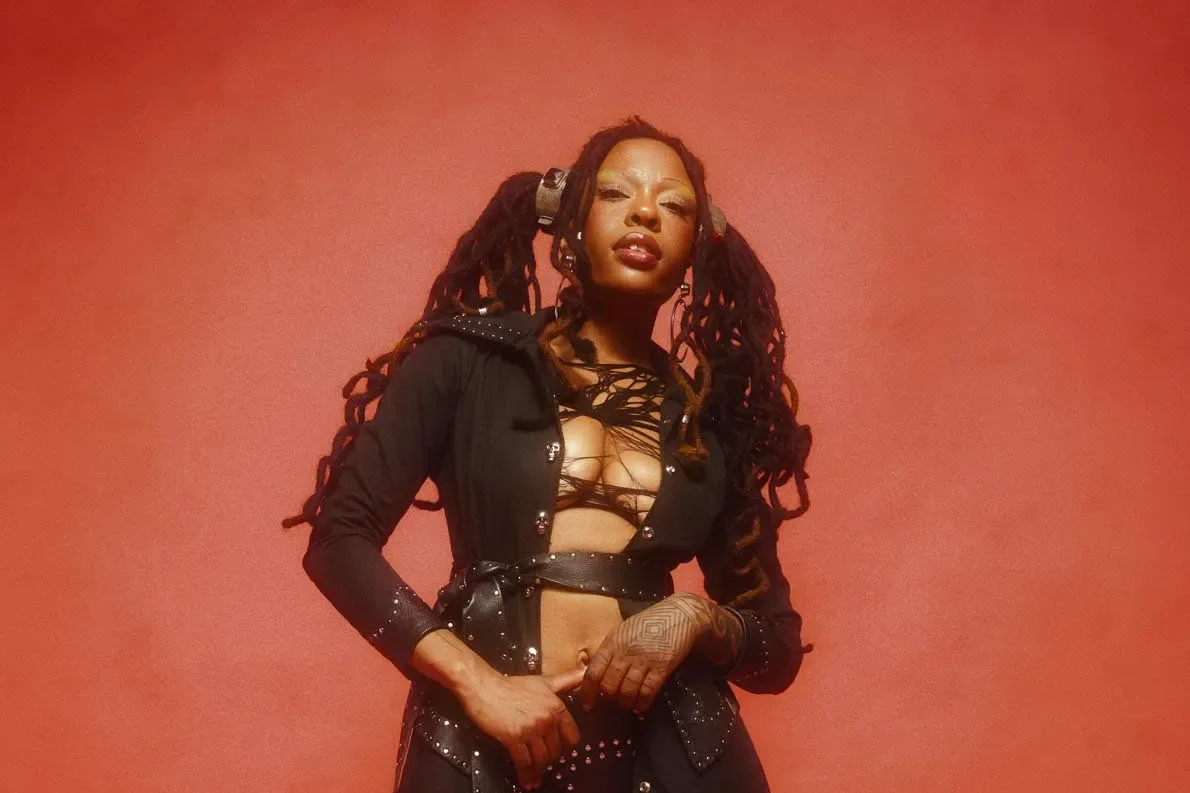 A woman with dreadlocks and tattoos wearing a black outfit, standing against a red background.