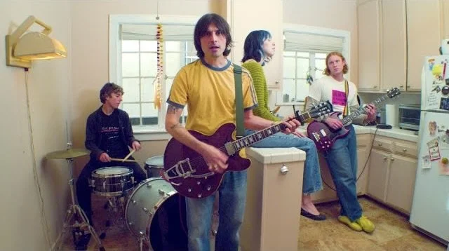 Four young adults playing music in a kitchen, one with a guitar, one with a drum, and others also with guitars.
