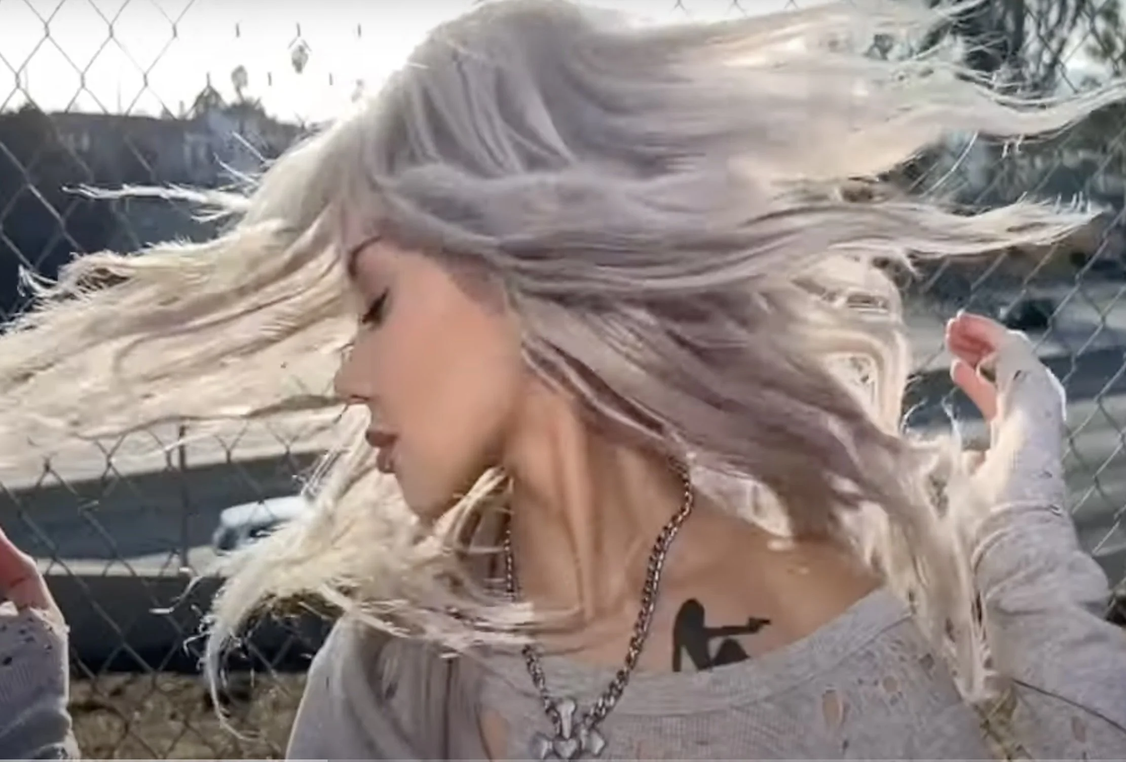 Fake Mink A woman with long blond hair leaning against a chain-link fence, with her eyes closed and head tilted downward, outdoors during daytime.