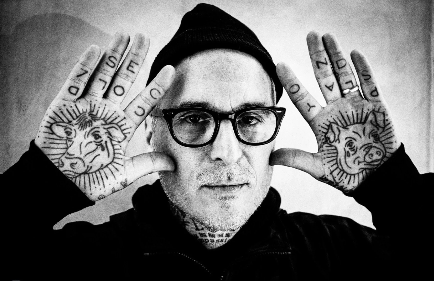Black and white photo of a man wearing glasses and a beanie, holding up his hands with tattoos of lions on his palms and the words 'DONT SEDUCE' on his fingers.