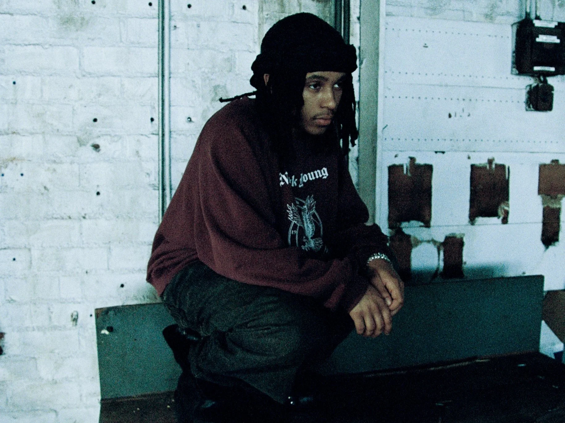 A young man with dreadlocks crouching against a white brick wall.