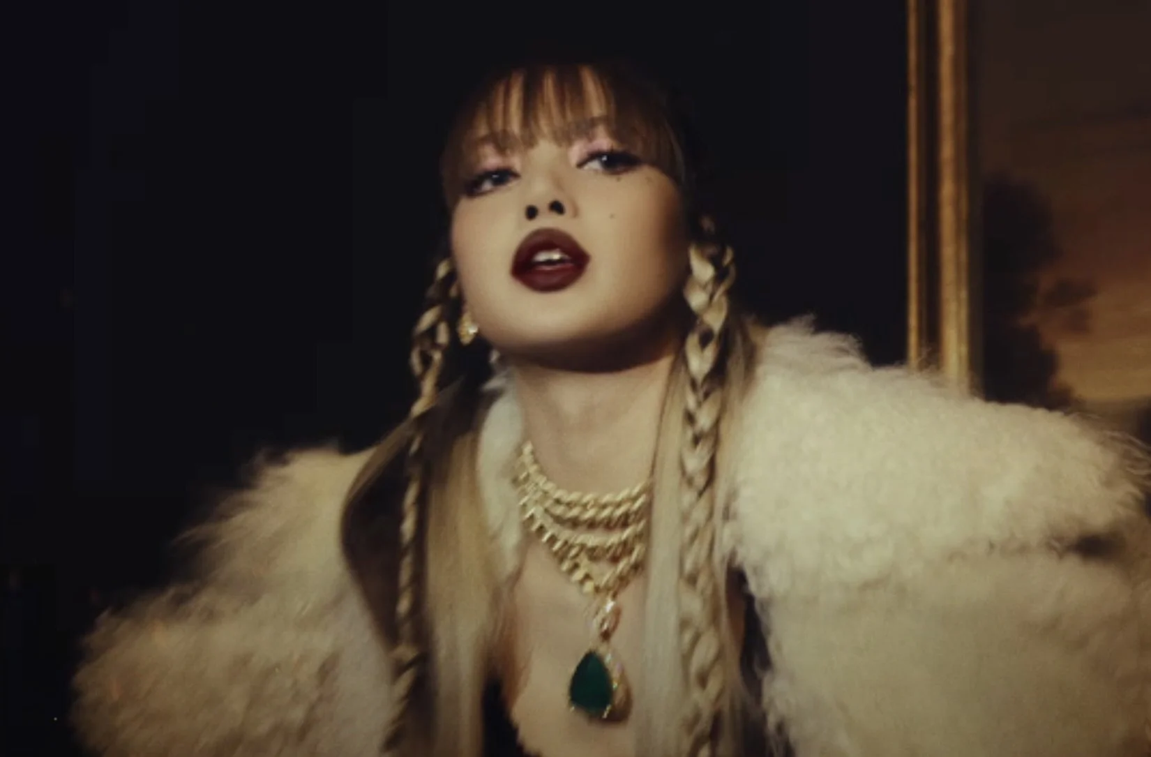 LISA Shows All Sides With New LP and Visual for “FXCK UP THE WORLD (Vixi Solo Version) A woman with long hair styled in braids, wearing bold makeup with dark lipstick, jewelry, and a furry coat, posing in a dark setting.