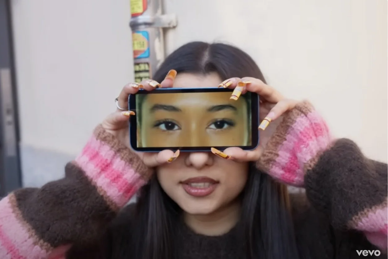 A woman with long dark hair holding a smartphone horizontally in front of her face, displaying a zoomed-in image of her eyes on the phone screen. She is wearing a multicolored sweater with pink, brown, and beige stripes and has brightly colored, striped nail polish.