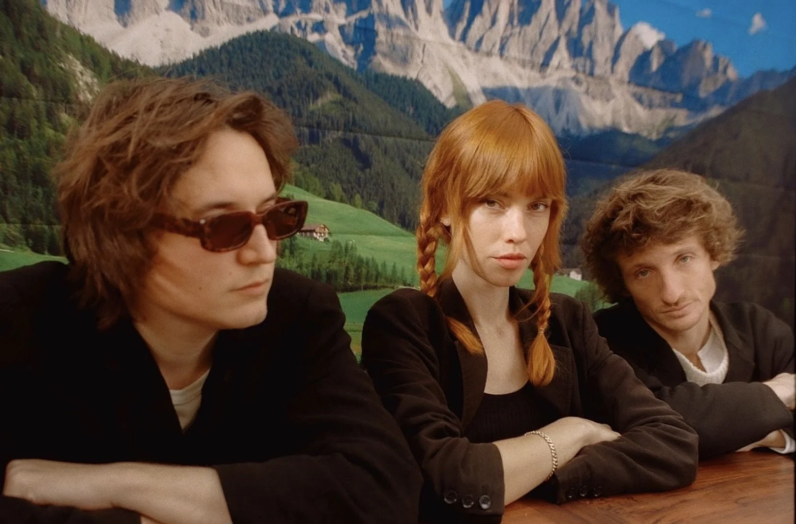 Oracle Sisters French Indie Three young adults sitting at a table with a mountainous landscape background, two with curly hair and one with straight red hair in braids, all with serious expressions.