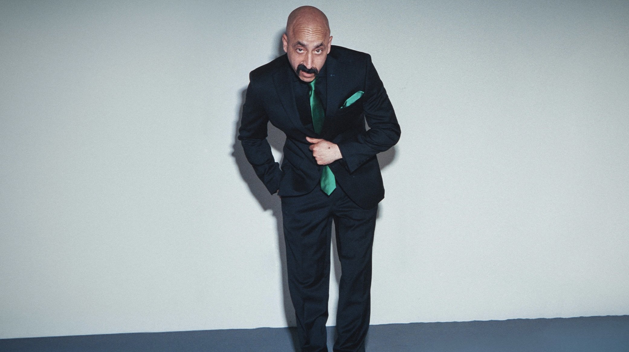 A man with a bald head, mustache, and beard, wearing a dark suit with a green tie and pocket square, is standing against a plain white wall, looking towards the camera, with a playful expression.
