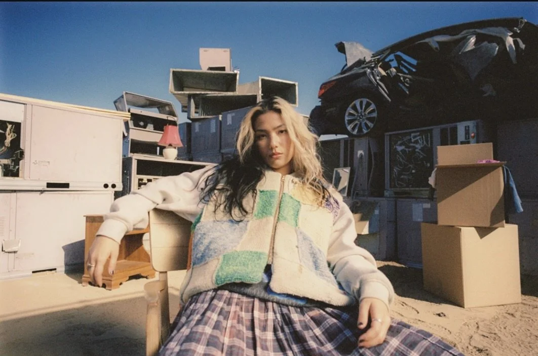 A young woman sitting outdoors with a relaxed expression, surrounded by stacked and disorganized household appliances, furniture, and a car on a trailer in the background, under a clear blue sky.