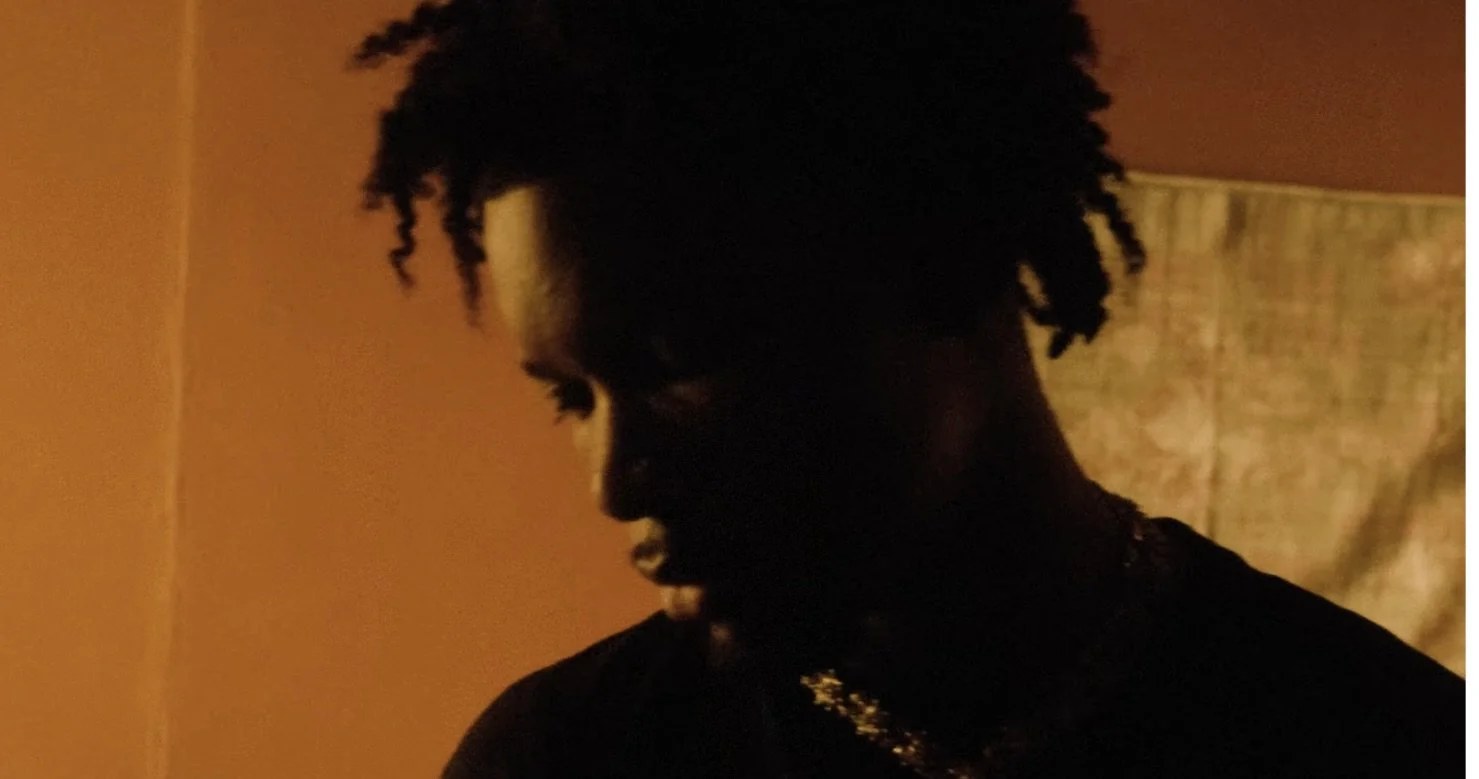 A side profile of a person with dark skin and natural curly hair, in a dimly lit room with orange walls.