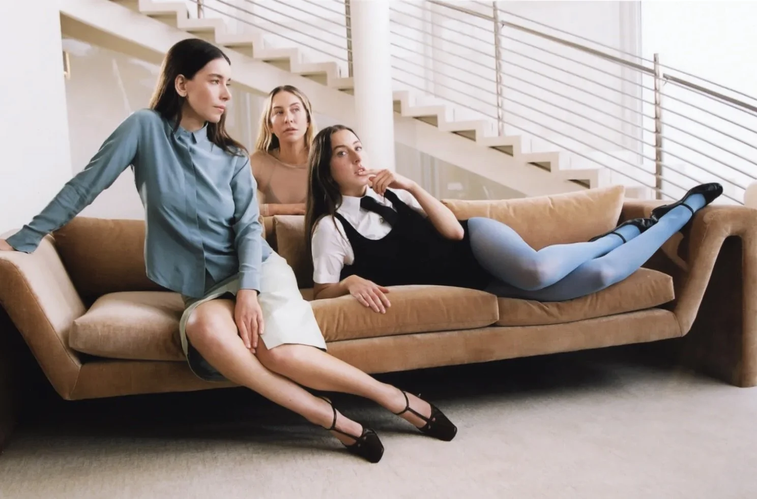 HAIM Three women on a beige couch in a modern living room. Two are sitting, and one is lying down with her legs extended across the couch. They appear to be posing thoughtfully, with a staircase and railing in the background.
