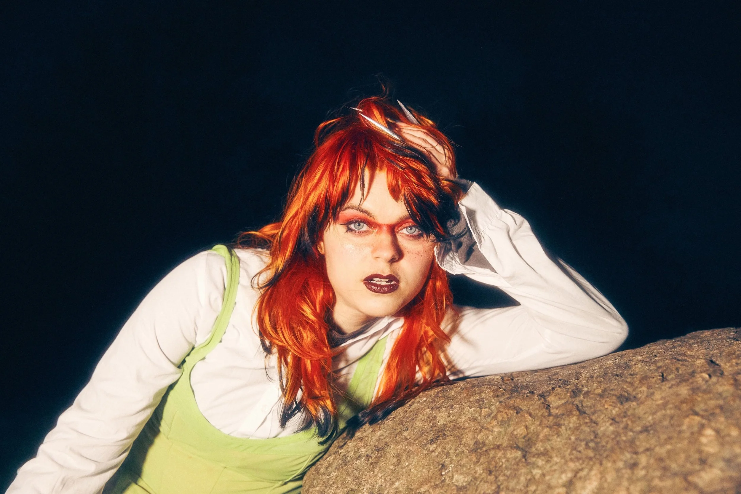 Ellie Dixon A woman with bright red hair and dark makeup leans on a large rock at night, wearing a white long-sleeve shirt and a light green dress.