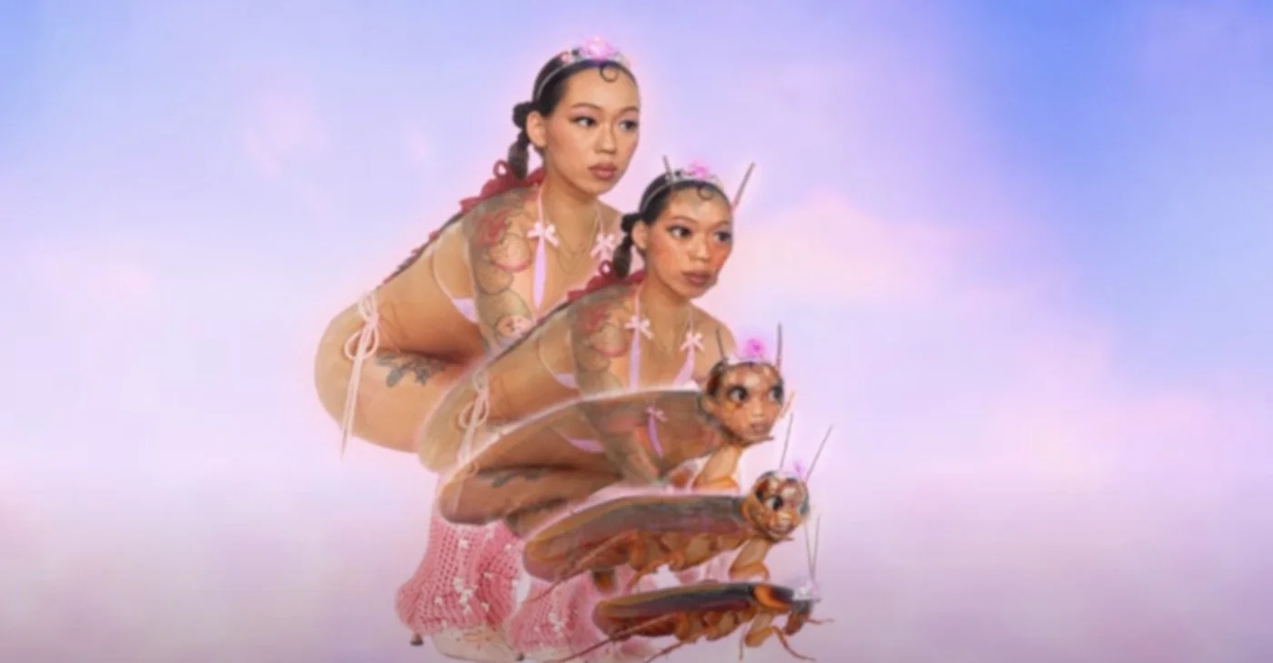 Multiple women with butterfly wings and and tattoos, viewed with a surreal, giant bug-like head, against a pastel-colored sky.
