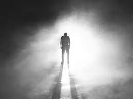 A solitary person walking down a foggy, deserted road with a bright light in the distance.