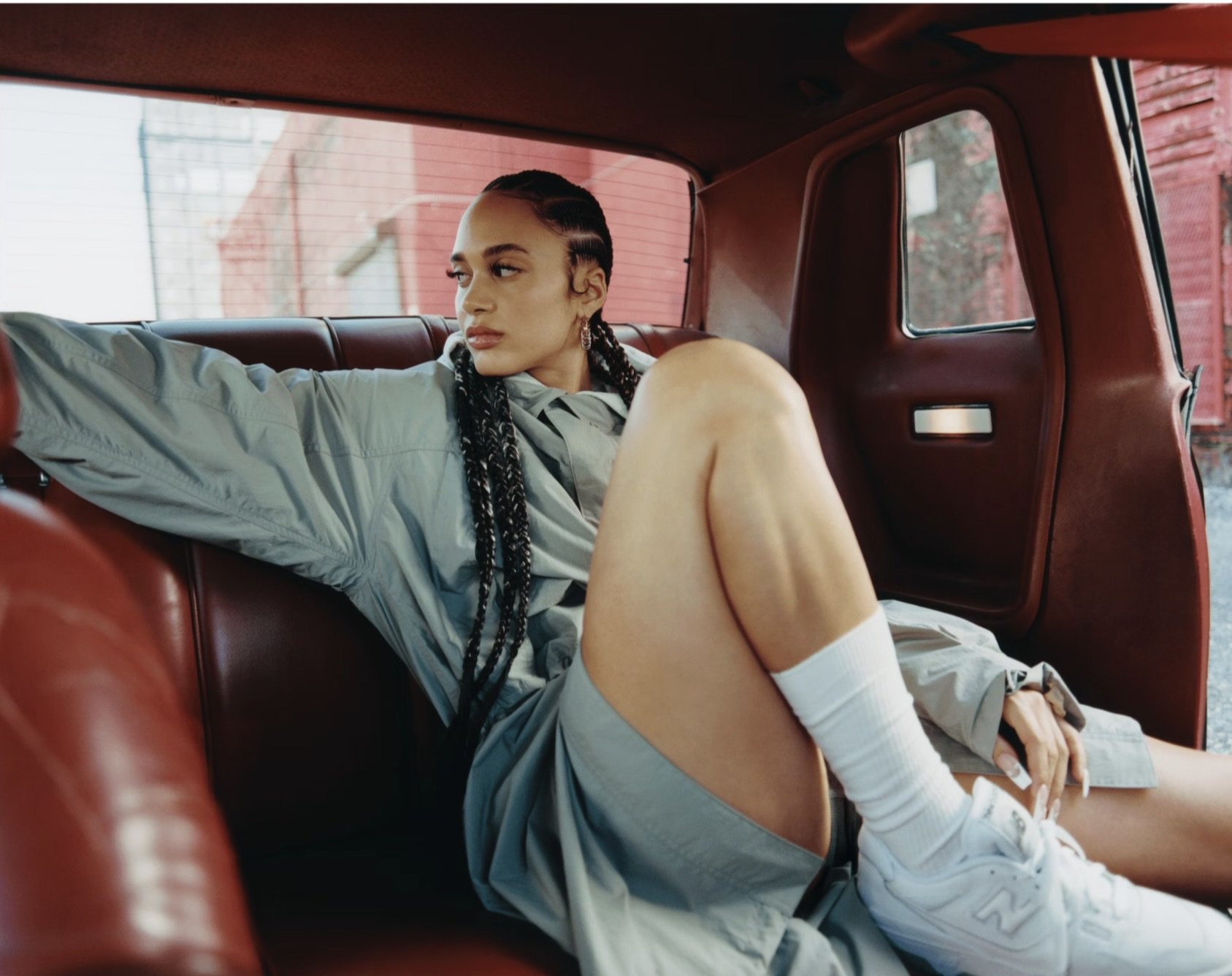 A woman with braided hair sitting in the back of a red vintage car, looking to her left with her arm stretched out on the seat.