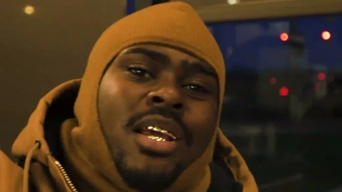 Close-up of a man wearing a brown beanie and hoodie, with a dark background and some blurry red and white lights.