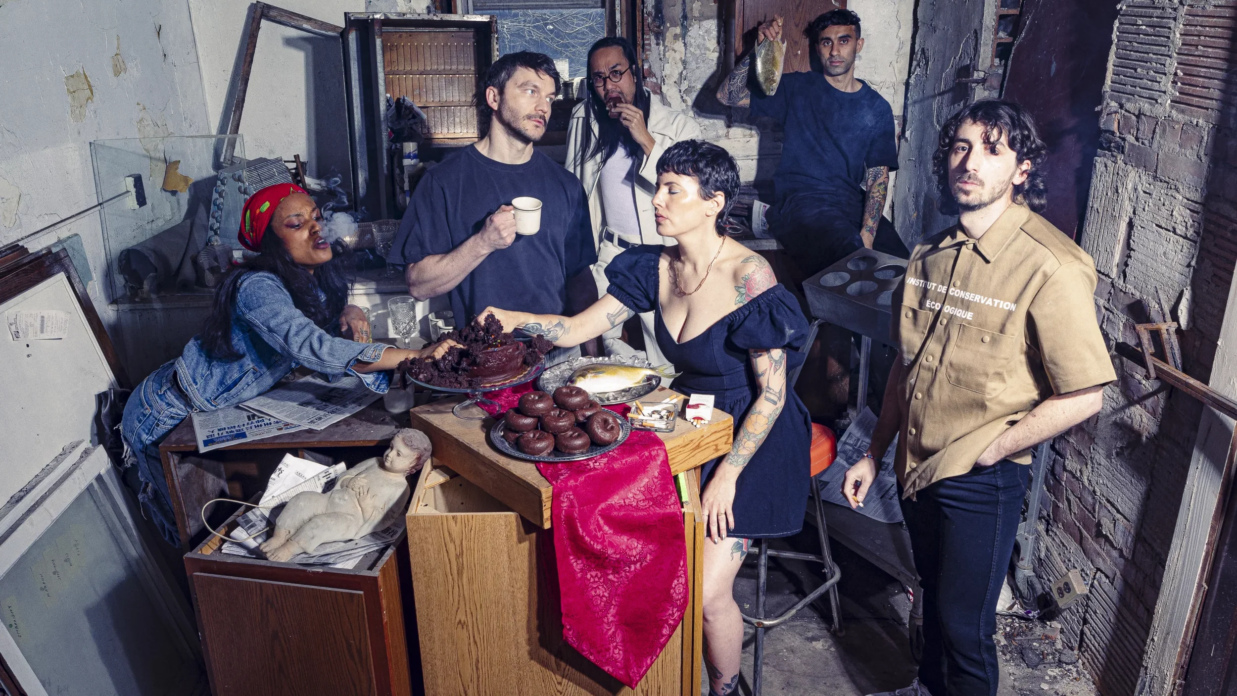 A group of six diverse adults gathered in a cluttered, industrial-style room with exposed brick walls and peeling paint, around a table filled with cakes, donuts, and snacks. One woman reaches for a piece of cake while a man with tattoos and short hair offers her a cupcake, another woman in a dress is seated and receiving a hug. Two men stand behind, one holding a cup and the other with a bag, while a woman wearing a headscarf leans over the table. The room has a mixture of vintage and modern elements, with miscellaneous items and scattered objects