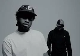 Two men standing in front of a plain background, both wearing dark caps and casual clothing.