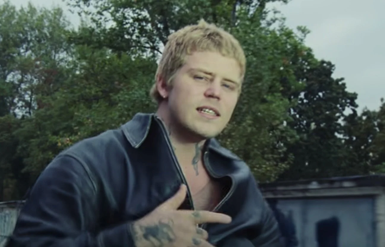 Yung Lean oung man with blond hair and tattoos on his fingers, wearing a black leather jacket, holding his chest, outdoors with trees in the background.