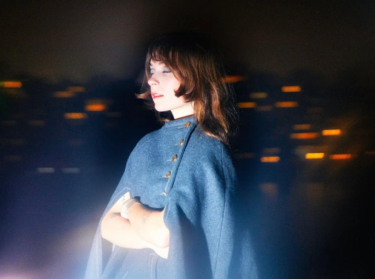 A woman standing outside at night wearing a dark blue denim jacket, with blurred city lights in the background.