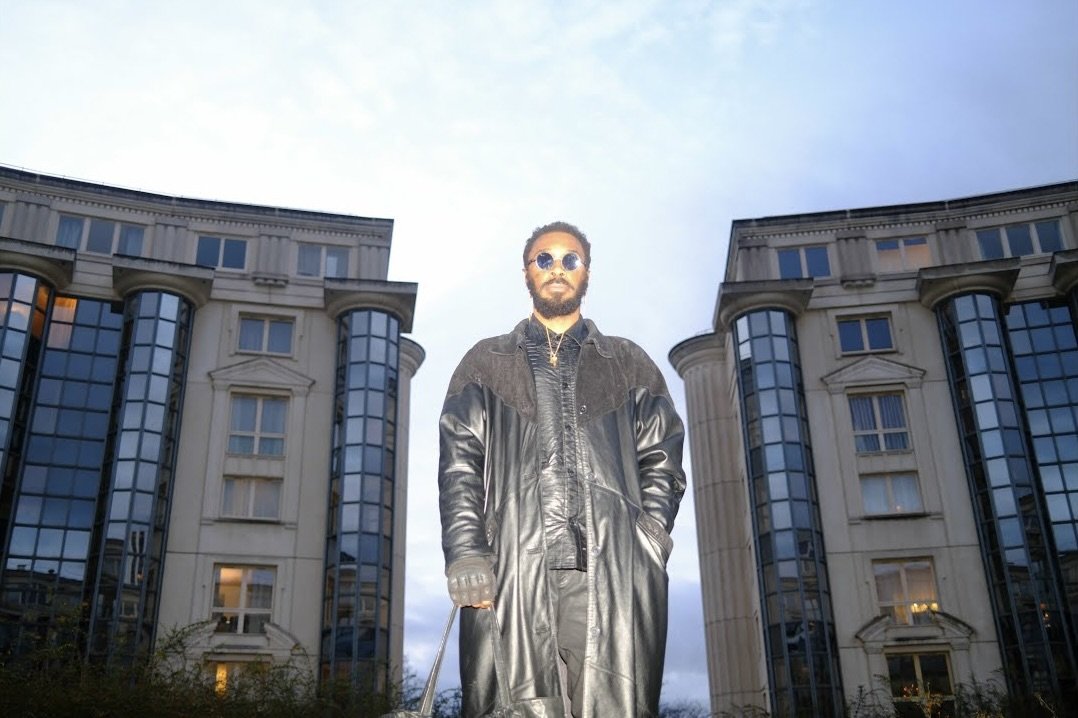 J PEG MAFIA A man stands outdoors in front of two large, modern buildings with glass windows, wearing sunglasses and a black leather coat.