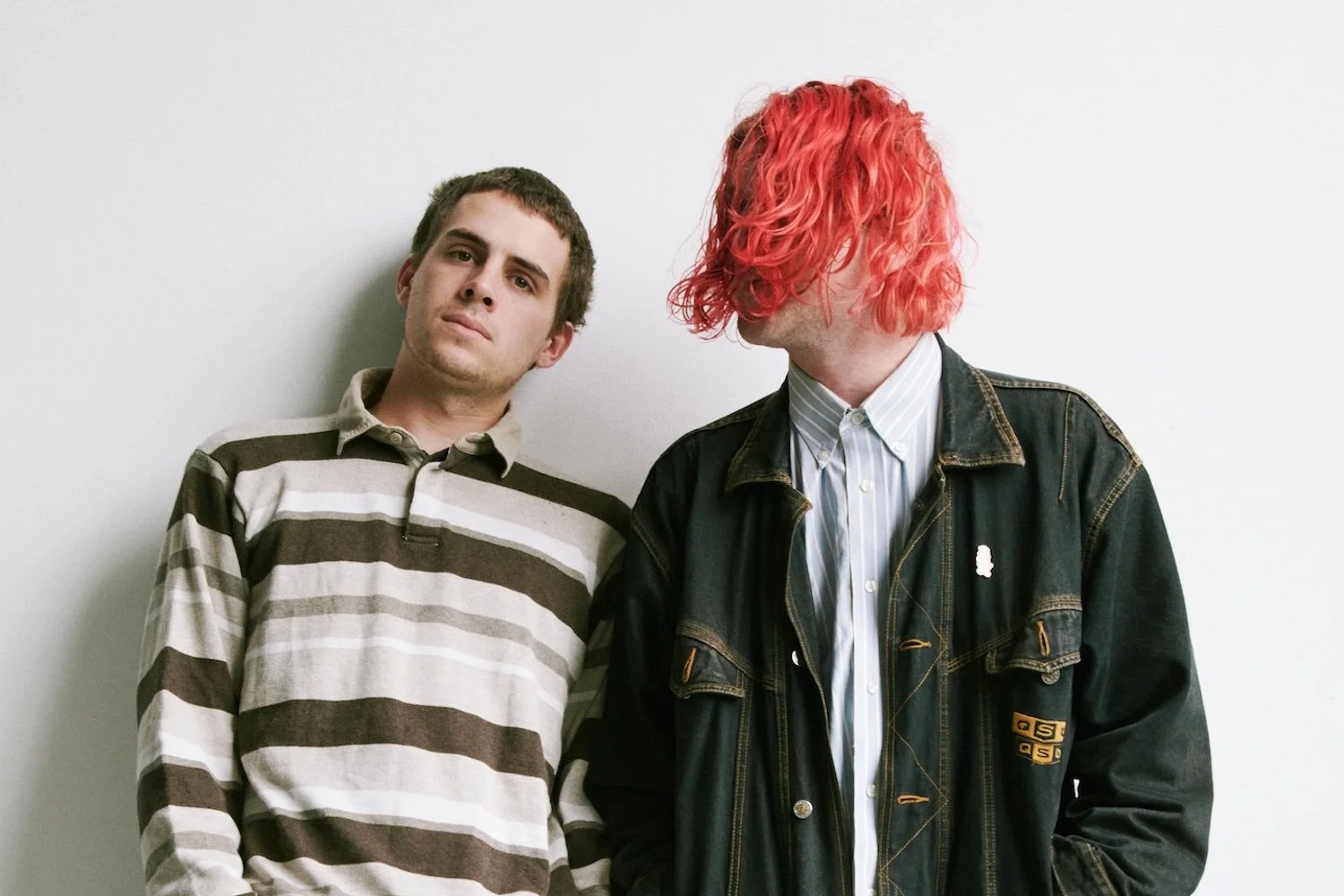 Two young men standing against a plain white wall, one with brown hair and a striped shirt, the other with bright pink hair and a denim jacket with patches.
