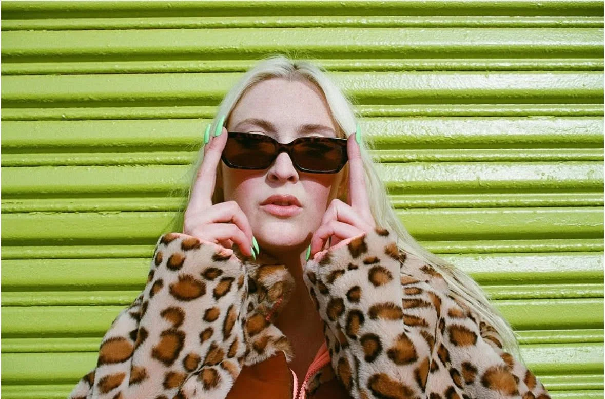 Woman with blonde hair wearing black sunglasses and a leopard-print coat, standing in front of a green corrugated metal wall.