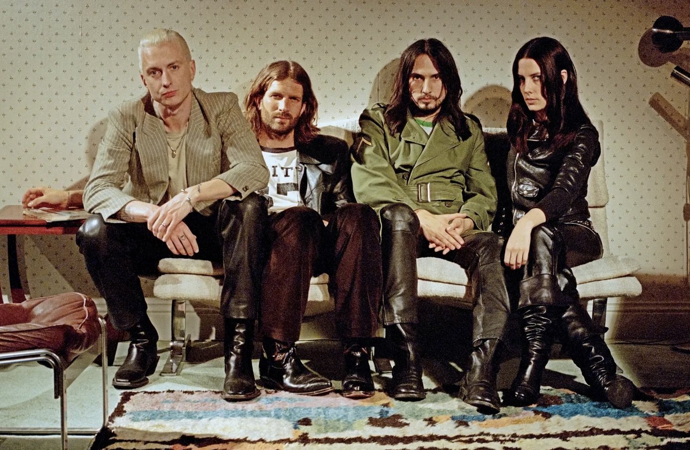 Four people sitting on a beige couch in a room with patterned wallpaper and a colorful rug.