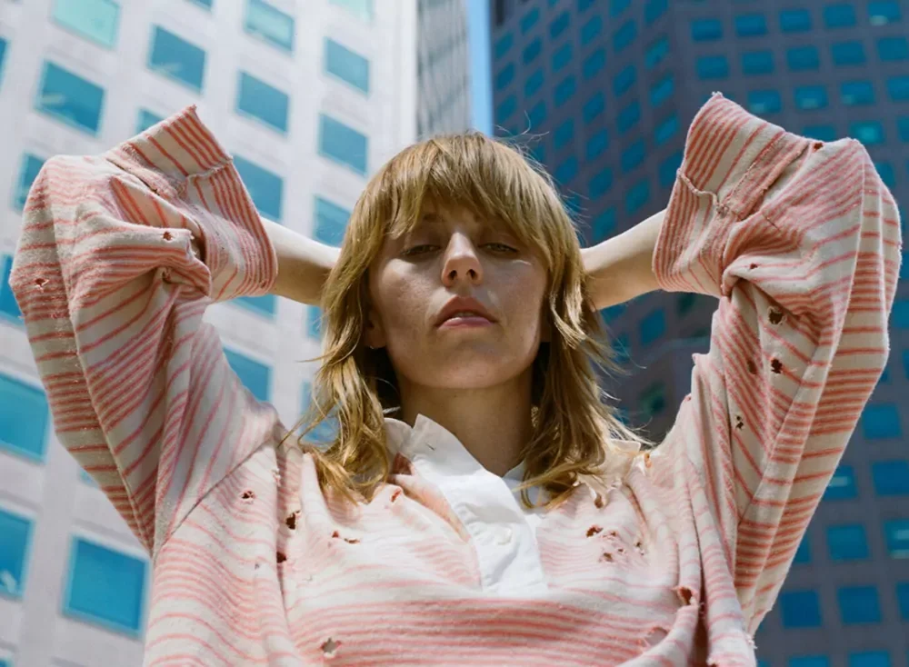 Young woman with light brown hair wearing a torn pink striped sweatshirt, standing outdoors in front of tall office buildings, with arms raised behind her head.