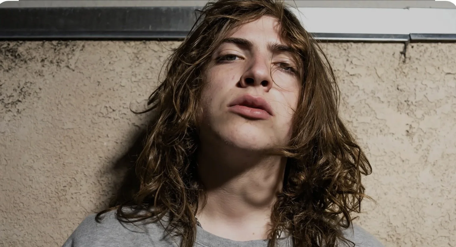 Close-up of a young person with long, wavy hair leaning against a textured wall, looking directly at the camera with a neutral expression.
