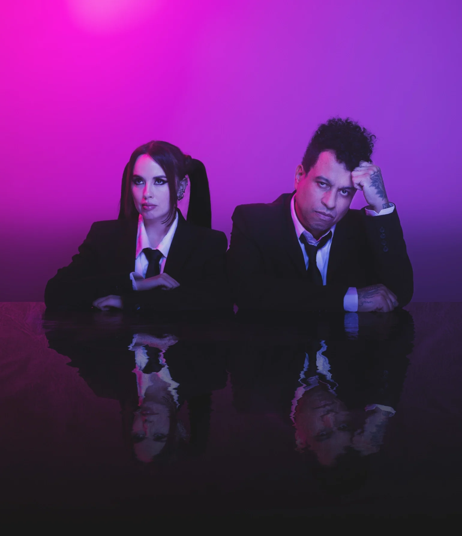 A man and a woman wearing black suits with white shirts sitting at a reflective dark table against a purple-pink gradient background.