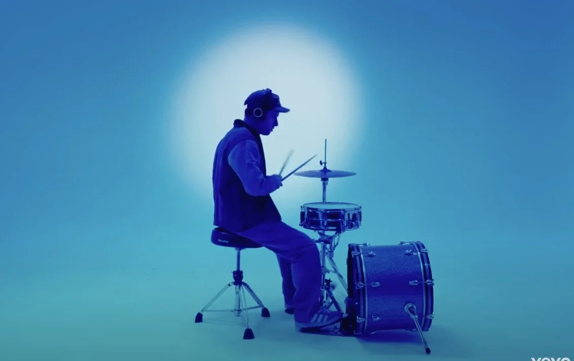 Luke Titus Silhouette of a person playing drums in front of a blue background with a bright light behind.
