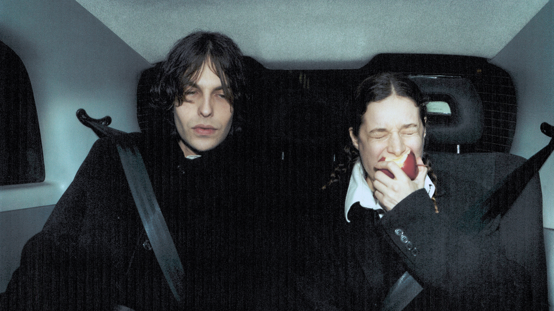 A young man with dark, wavy hair and pale skin sitting in the backseat of a car wearing a black outfit, and a young woman with dark hair in braids and light skin, grinning while biting into a red apple, also in the car.