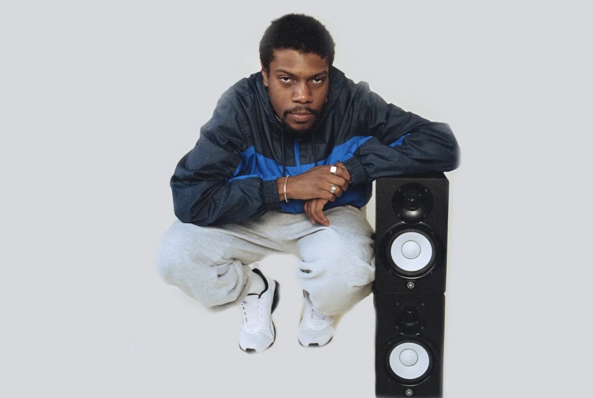 A young man crouching next to a tall black speaker with white accents, wearing a navy blue and black windbreaker, gray sweatpants, and white sneakers, with a serious expression.