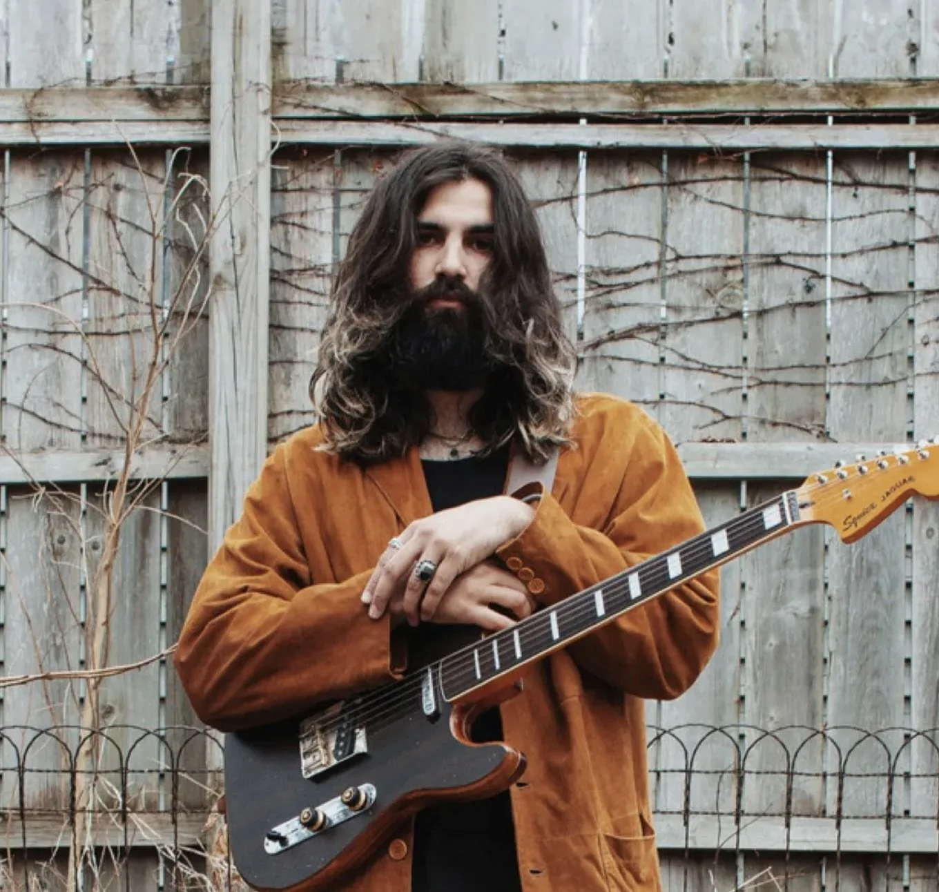 A man with long, wavy hair and a full beard standing outdoors in front of a wooden fence. He is wearing a brown jacket and holding an electric guitar.