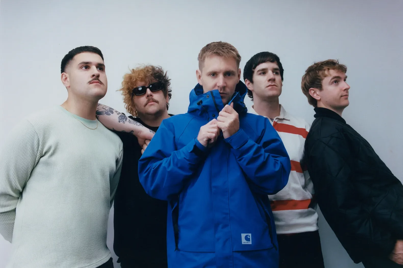 Five young men standing in a line against a plain white wall, with serious expressions. The man in the center is wearing a bright blue jacket and holding it close to his chin. The others wear casual clothing, including a white sweater, a black shirt with tattoos, sunglasses, a striped shirt, and a black jacket.