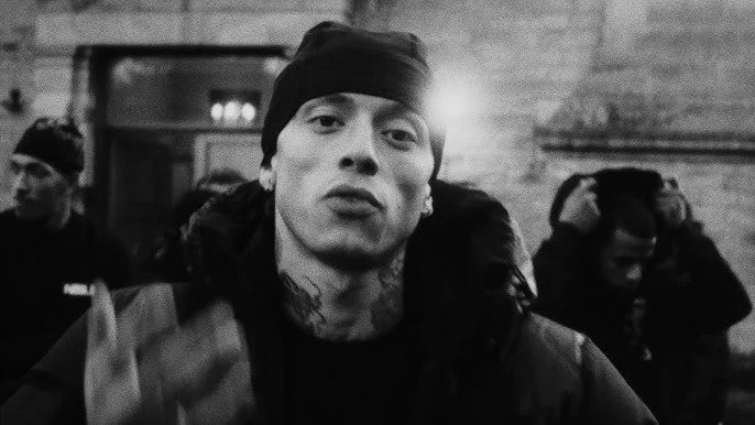Central Cee Black and white photo of a woman with tattoos and earrings, wearing a headband and jacket, making a gesture towards the camera, with a person in the background adjusting headphones.