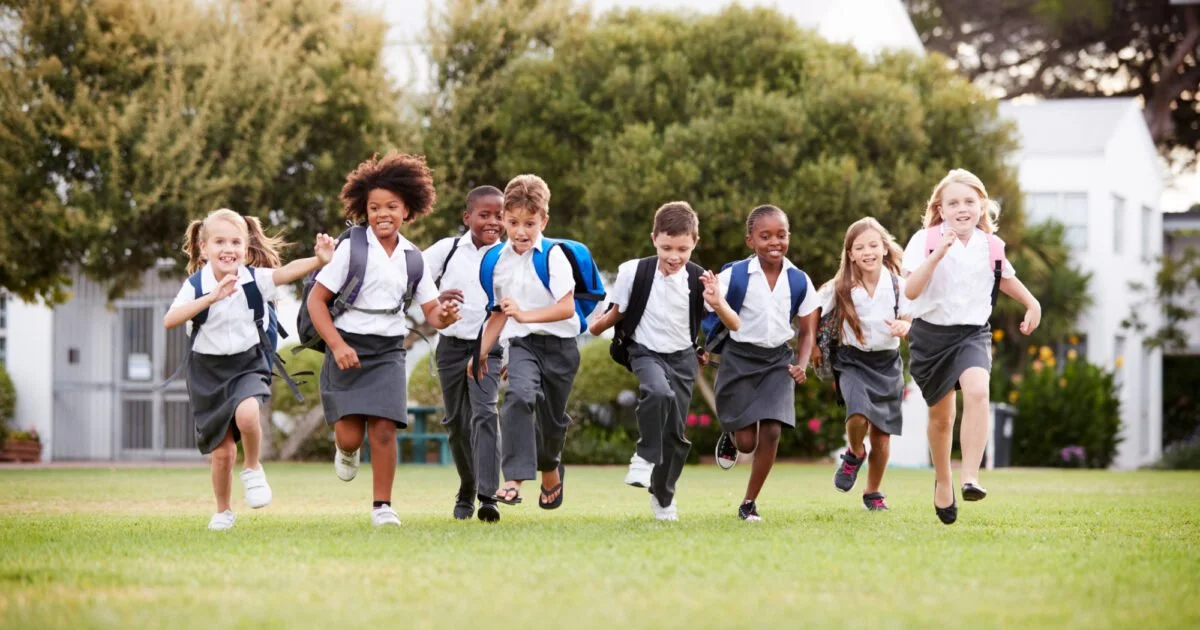 A group of children in school uniforms running on a grassy field, carrying backpacks.